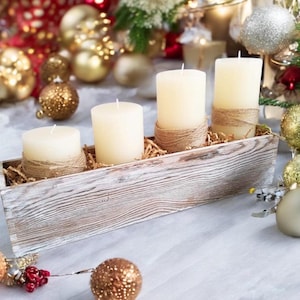 May include: Four white pillar candles in a rustic wooden box with a natural twine wrap. The box is filled with shredded paper and the candles are arranged in a row. The box is sitting on a white surface.