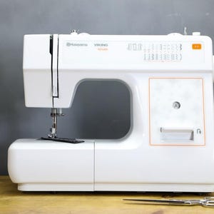 Op de afbeelding: Een witte Husqvarna Viking E10 naaimachine met een zilveren naald en zwarte naaldplaat. De machine heeft een oranje accent en een bedieningspaneel met verschillende steekopties. Een schaar en garenklosjes staan op de voorgrond.