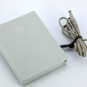May include: A rectangular, light grey foot pedal controller with a textured surface. A long, light grey power cord is attached to the side of the pedal, coiled and secured with a black plastic cable tie, ending in a small cylindrical power connector.