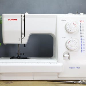 Op de afbeelding: Witte Janome naaimachine, Model 7021, met meerdere knoppen en instellingen. De machine heeft een naald, een naaivoet en een werkoppervlak. Een schaar en garenklosjes staan op de voorgrond.