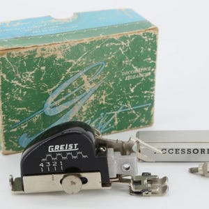 May include: A black Greist decorative zigzag sewing machine attachment with a numbered dial from 1 to 4, a silver metal presser foot, and a small screw. It is accompanied by a green and blue cardboard box and a silver box labeled ACCESSORIES.