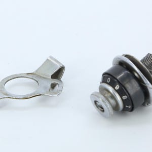 Könnte beinhalten: Ein zylindrischer, schwarz-silberner Drehknopf mit einer nummerierten Skala von null bis neun, ausgestattet mit einem Gewindeschaft aus Metall und einer separaten silberfarbenen Metallhalterung mit runder Öffnung und gebogener Lasche.