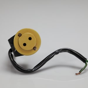 May include: A vintage-style electrical socket featuring a circular, mustard-yellow faceplate with two pin holes and two small screws. A black insulated electrical cord extends from the back, terminating in two exposed, stripped copper wire ends.
