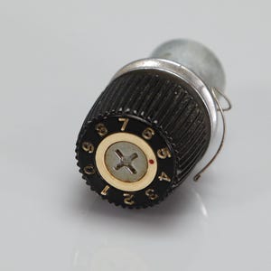 May include: A black cylindrical control knob with vertical ridges and a silver center featuring a cross-head screw. The top edge displays white numbers from zero to nine in a circular pattern, with a small red dot indicator near the number five.