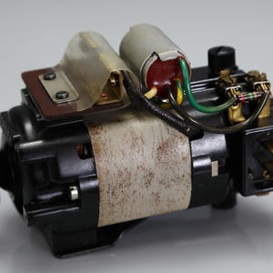 May include: A black electric motor featuring a metallic pulley on one end, a cylindrical capacitor with green and black wiring, a brown metal mounting bracket, and a textured beige band wrapped around the central housing with visible rust spots.