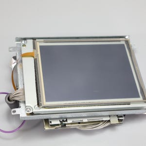 May include: A square liquid crystal display screen module featuring a dark gray panel mounted on a silver metal frame. The component includes a thin purple wire, a gray ribbon cable, and a small orange flexible circuit connector attached to the side.