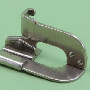 May include: A silver-coloured metal sewing machine presser foot attachment featuring a U-shaped frame, a hinged joint with a small screw, and a guide channel. The component is constructed from polished steel with visible rivets and a hook-like top.