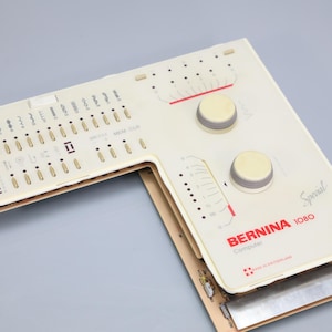 May include: An off-white Bernina 1080 Computer Special sewing machine faceplate featuring a L-shaped control panel with numerous stitch selection buttons, two circular adjustment knobs, red text branding, and visible internal electronic circuitry.