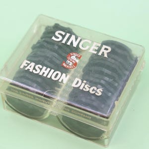 May include: A clear plastic storage case containing black Singer Fashion Discs for sewing machines. The lid features the white text SINGER FASHION DISCS and a red circular logo with the text SINGER SEWING MACHINES. The base is marked with THE SINGER MANUFACTURING COMPANY.