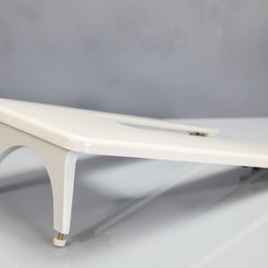 May include: A white slanted laptop stand with a smooth, glossy finish. The stand features a raised platform supported by two arched legs at the front, each equipped with small, adjustable feet for stability and a slight incline for ergonomic viewing.