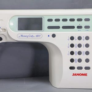 Könnte beinhalten: Eine weiße Kunststoff-Frontplatte für die Janome Memory Craft 6600 Nähmaschine mit einem digitalen Anzeigebereich, einer Reihe rechteckiger Tastenöffnungen, einem Schiebeschalter und einem Raster aus kreisförmigen Öffnungen mit blauen Stichsymbolen.