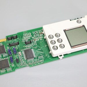 May include: A green printed circuit board featuring a white plastic module with a square LCD screen and six circular buttons. The board includes various electronic components, integrated circuits, and a gold-plated edge connector labeled 2027.