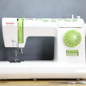 Op de afbeelding: Een witte Toyota naaimachine met groene accenten. De machine heeft een groene draaiknop, een naald en het woord "TOYOTA" in het rood. De machine heeft een bloemenpatroon en staat op een houten oppervlak.
