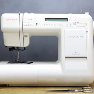 Op de afbeelding: Een witte Janome Memory Craft 6000 naaimachine met een digitaal display en bedieningsknoppen. De machine staat op een houten oppervlak met een schaar en klosjes garen. De merknaam is zichtbaar.