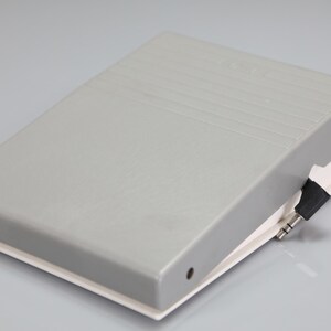 May include: A rectangular gray plastic foot pedal controller with a white base and a black audio jack connector attached by a short cord. The top surface of the pedal features a series of horizontal embossed lines for grip and a small circular hole.