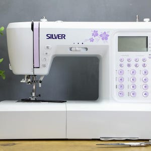 Op de afbeelding: Een witte Silver naaimachine met een digitaal display en lavendelkleurige accenten. De machine heeft genummerde knoppen en bloemmotieven. Een schaar en garenklosjes liggen op de voorgrond.