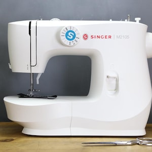 Op de afbeelding: Een witte Singer M2105 naaimachine met een zwarte naald en draad. De machine heeft een ronde draaiknop met het Singer-logo en modelnummer. Schaar en garenklosjes op de voorgrond.