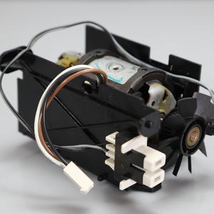 May include: An electric motor assembly featuring a cylindrical metal housing, a black plastic frame, and a small black cooling fan. The unit includes a white plastic connector block with two ports and several thin wires in black, white, and brown.