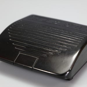 May include: A black plastic sewing machine foot pedal controller with a rectangular shape, featuring a series of horizontal raised ridges across the top surface for grip and a small rectangular indentation at the front edge for operation.