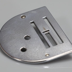 Op de afbeelding: Een zilverkleurige metalen naaimachine steekplaat met een halfronde rand aan de ene kant en een rechte rand aan de andere kant. De plaat bevat twee ronde schroefgaten en twee parallelle rechthoekige sleuven voor de naald en transporteur.