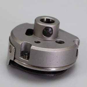 May include: A metallic silver sewing machine bobbin case featuring a cylindrical center post with a black slotted screw, a circular body with cutouts, and a small tension spring mechanism visible on the side of the circular metal housing.