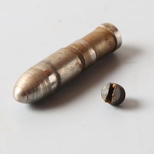 Könnte beinhalten: Eine zylindrische Metallkomponente mit abgerundeter Spitze und einer verwitterten, angelaufenen Oberfläche, neben einem kleinen, dunklen, kreisförmigen Metallschraubenkopf mit einem zentralen Schlitz für einen Schlitzschraubendreher.