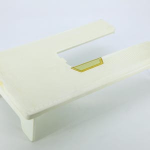 May include: A white plastic sewing machine extension table with a rectangular cutout. The surface features a grid pattern and a small, centred yellow rectangular accent piece. The table is supported by two legs on one side and a single leg on the other.