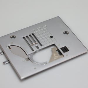 May include: A rectangular silver-coloured metal sewing machine needle plate featuring a grid pattern with markings 10, 20, and 30. The plate includes a central needle slot, a feed dog opening, a bobbin case area, and two circular screw holes.