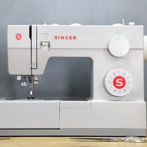 Op de afbeelding: Een witte Singer Heavy Duty naaimachine met rode accenten. De machine heeft meerdere knoppen en een naald. Schaar en garenklosjes staan op de voorgrond. Een groene plant staat op de achtergrond.