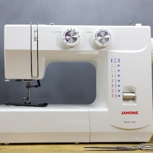 Op de afbeelding: Een witte Janome Model 1560 naaimachine met zilveren knoppen en een naald. De machine heeft een selectie van steekpatronen aangegeven met blauwe en rode symbolen. Een schaar en spoelen garen staan op de voorgrond.
