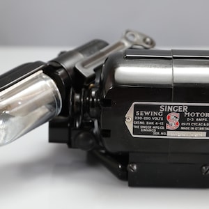 May include: A black Singer sewing motor with an attached light fixture. The metal identification plate reads: SINGER SEWING MOTOR, 230-250 VOLTS, 0-3 AMPS, CAT.NO. BAK 4-12, 25-75 CYC.AC & DC, THE SINGER MFG.CO. SIMANCO, MADE IN GT.BRITAIN.
