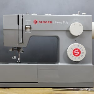 Op de afbeelding: Een grijze Singer Heavy Duty naaimachine met witte wijzerplaten en rode accenten. De machine heeft een naald, draad en een werkoppervlak. Spoelen garen en een schaar zijn ook zichtbaar.
