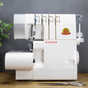 Può includere: Una taglia e cuci Singer bianca con il marchio in rosso. La macchina ha più rocchetti di filo e un paio di forbici. Sullo sfondo c'è una pianta in un vaso nero.