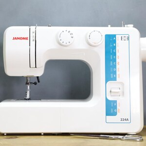 Op de afbeelding: Een witte Janome 224A naaimachine met een zilveren naald en een blauw-wit bedieningspaneel. De machine heeft twee witte draaiknoppen en de merknaam "JANOME" in het rood. Een schaar en spoelen garen staan op de voorgrond.