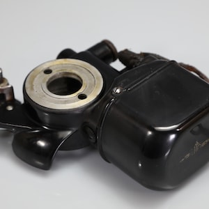 May include: A vintage black metal mechanical component featuring a circular silver-toned opening, a lever mechanism, and a rectangular housing with the word "Luthor" printed in gold script. The device includes attached frayed electrical wiring.