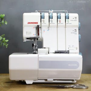 Könnte beinhalten: Eine weiße Janome 6234XL Overlock-Nähmaschine mit vier blauen Spannungsreglern an der Oberseite, einem Nadelbereich mit Metallnähfuß und einer Frontplatte mit der Modellnummer. Die Maschine hat ein sauberes und kompaktes Design.