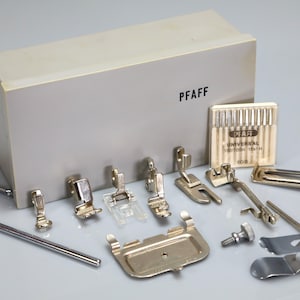 May include: A collection of silver-toned metal sewing machine presser feet and accessories arranged in front of a grey plastic storage case labeled PFAFF. Includes a pack of PFAFF Universal 130/705H 15x1H 80/12 needles, a metal guide bar, a screw, and various specialized feet.