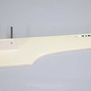 May include: An off-white sewing machine extension table with a smooth, flat surface and a curved edge. Two vertical metal pins are attached to the left side. A rectangular white plate with the black text VIKING is affixed to the right side.
