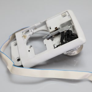 May include: A white plastic electronic component housing featuring an internal black mechanical lever mechanism and a flat, flexible ribbon cable extending outward, terminating in a small red plastic connector with multiple metal contact pins.