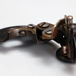 Pode incluir: Um acessório de máquina de costura de metal vintage com um braço preto curvo, uma pequena roda circular e vários parafusos prateados. O componente mecânico inclui um suporte de metal articulado e uma borda texturizada na roda.
