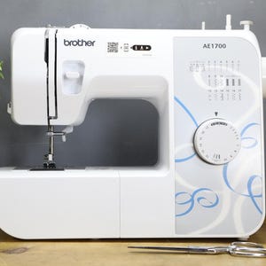 Op de afbeelding: Een witte Brother AE1700 naaimachine met blauwe en grijze accenten. De machine heeft een draaiknop, steekselectie-indicatoren en een naald. Een zilveren schaar en spoelen garen zijn ook zichtbaar.