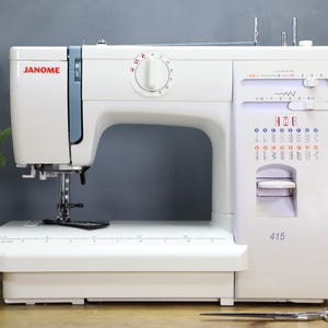 Op de afbeelding: Een witte Janome 415 naaimachine met blauwe en rode accenten. De machine heeft een draaiknop, een steekselectiepaneel en een naald. Een schaar en spoelen garen staan op de voorgrond.