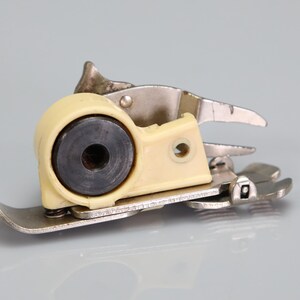 May include: A metal sewing machine presser foot attachment featuring a cream-colored plastic housing, a central black circular component with a hollow center, and a silver-toned metal base plate with a flat, elongated shape and a small screw.