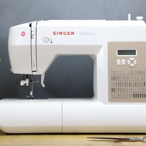 Op de afbeelding: Een witte Singer Brilliance naaimachine met rode merknaam. De machine heeft een digitaal display en diverse steekopties. Een schaar en garenklosjes liggen op een houten oppervlak.