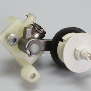 May include: A mechanical assembly featuring a translucent off-white plastic bracket, a silver metal screw, a coiled metal spring, a dark grey metallic pivot arm, and a black rubber ring attached to a white plastic circular component with a central pin.