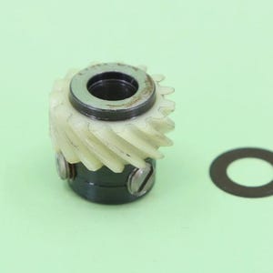 May include: A mechanical gear assembly featuring a white plastic helical gear mounted on a dark metal cylindrical base with two visible screws, accompanied by a separate thin, flat, dark metal circular washer positioned to the right side.