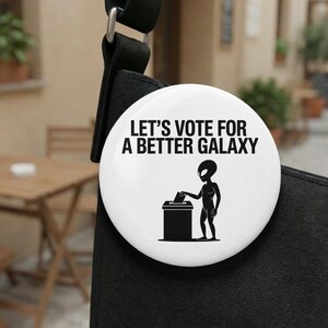 Op de afbeelding: Een witte knoop met de tekst "LET'S VOTE FOR A BETTER GALAXY" in het zwart. Een zwart silhouet van een alien plaatst een stembiljet in een stembus. De knoop is bevestigd aan een zwarte band.