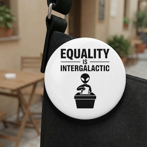 Puede incluir: Un botón blanco con el texto negro "EQUALITY IS INTERGALACTIC". Debajo, un gráfico en blanco y negro de una figura alienígena votando. El botón está sujeto a una bolsa negra.
