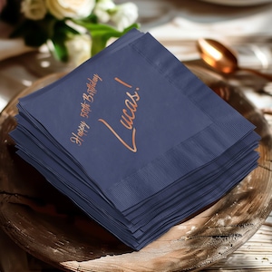 Può includere: Una pila di tovaglioli di carta blu navy con una scritta color rame. Il testo recita "Happy 50th Birthday Lucas!". I tovaglioli sono impilati su un piatto di legno, adatti per una festa di compleanno.