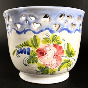 Peut inclure: Un cache-pot en céramique orné d'un motif floral peint à la main avec une grande rose rose, des feuilles vertes et des touches bleues. Le bord supérieur présente une découpe en forme de cœurs et une bordure bleu clair sur fond crème.
